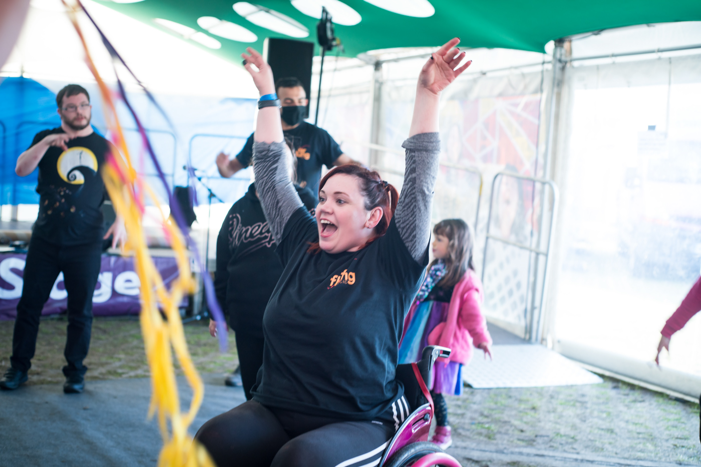 Hannah is in the centre of the image - Hannah is sitting in her manual wheelchair with bright pink wheels.  She is a white female, has brown hair tied half up, and is wearing a black T shirt with an orange and red Far Flung logo.  She is teaching a workshop in a festival marquee, and is stretching up to the ceiling with a huge smile on her face. In the background children and adults are dancing and in the foreground are colourful yellow, pink and purple ribbons sweeping across the front of the image.